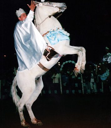 Cavaleiros da Cavalhada em formação na praça, diante da Matriz iluminada, à noite.