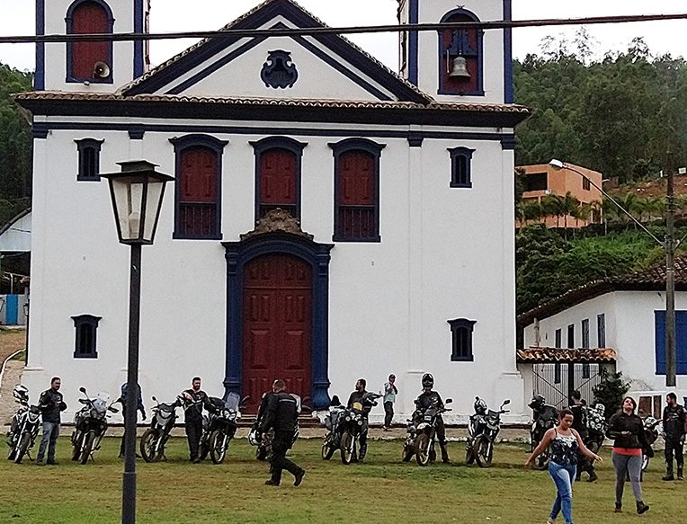 Trilha de terra subindo entre a vegetação, com as serras ao fundo.
