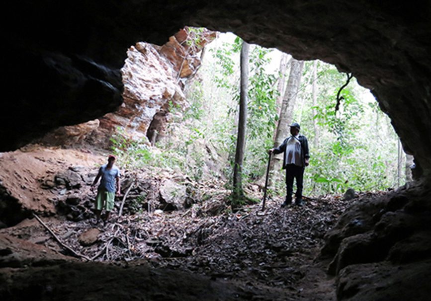 Entrada escura de galeria de mina escavada em barranco de terra vermelha, cercada de vegetação.
