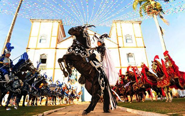 Cavalo branco empinado diante da igreja iluminada à noite, montado por cavaleiro de azul, com fileiras de cavaleiros ao fundo.