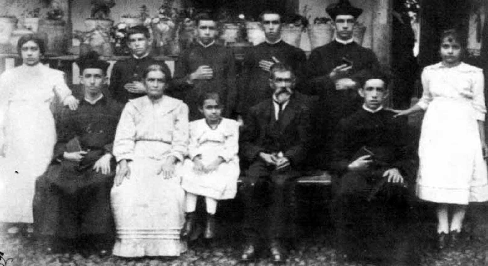 Retrato de grupo em preto e branco: casal sentado ao centro com uma criança, cercado por mulheres de vestido claro e seis homens jovens de batina.