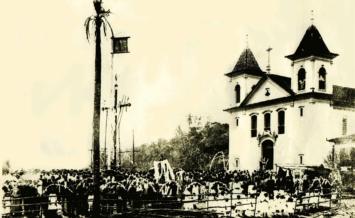 Fotografia antiga em preto e branco da Matriz de duas torres, com multidão reunida no largo em frente e um mastro alto à esquerda.